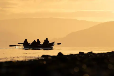 Anouk Schurink Silhouette of a rowing boat with five people in it, and oars out to the side, with some water splashing, in early evening light, with land of different shades in the foreground and background.