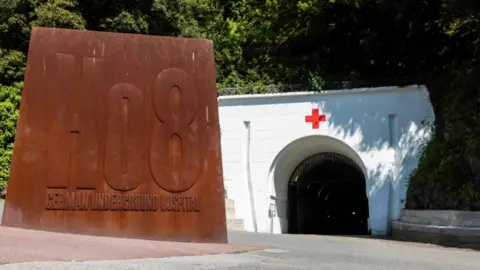 The entrance to Jersey War Tunnels. There is a large white stone archway with a red cross painted above the tunnel entrance. In front is a large rusted metal sign saying 'Ho8 German underground hospital'