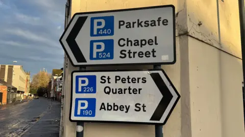 Two street signs mounted on a narrow corner building pointing to four different car parks - two in each direction.