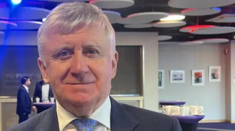 BBC Joe Moynihan is wearing a blue suit and a light blue tie. He has white hair. 