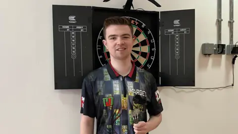 Jenson, with darts in his left hang, looks towards the camera in front of a dartboard. He has short dark hair and wears a black top with a slight chequered pattern with various images.