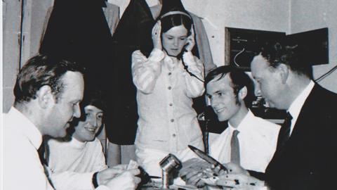 Four men sat down in front of mics with one woman stood up with headphones on - pic in black and white