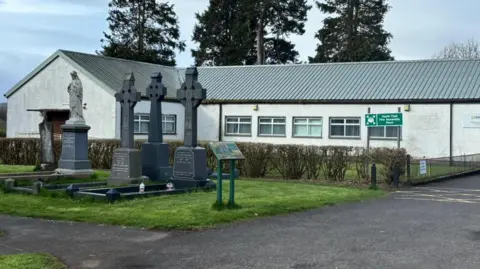 The image shows Claudy youth club, a white building with a tin roof. It is set behind a cemetery. A number of gravestones can be seen in the foreground