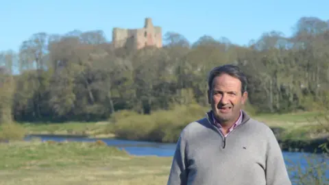 Northumberland County Councillor Patrick Lambert. He has short salt and pepper hair and is wearing a grey jumper. He is standing in an open area surrounded by grass, with a stream and a castle ruin in the background.