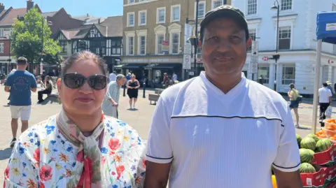 Uxbridge shoppers Piyara on the right wearing a white top and his wife wearing sunglasses, a floral top and chiffon floral scarf around her neck. In the background there is Uxbridge town centre square and part of a fruit stall displaying watermelons.