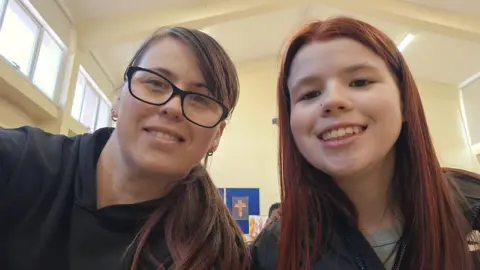 Lillie-Mae is pictured with her mum Katie. Katie is on the left of the picture, she has long brown hair swept over her left eye and is wearing glasses with black frames. Lilly-Mae in on the right. She has red hair and is smiling at the camera.