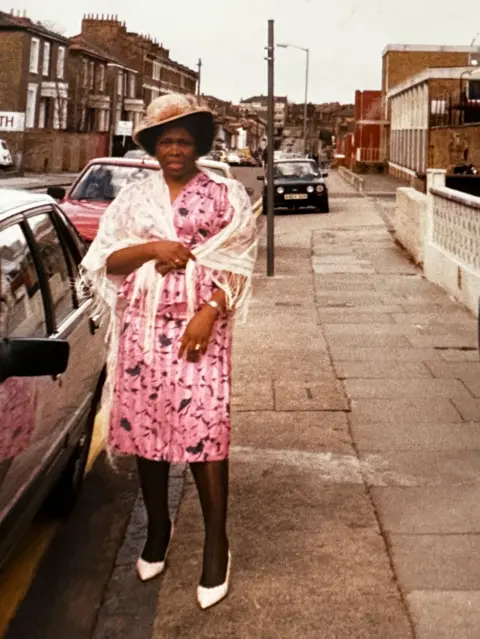 Jessie Reid Jessie wearing a pink dress and white shawl, white heels, standing on a residential street. She also wears a hat decorated with pink flowers. 