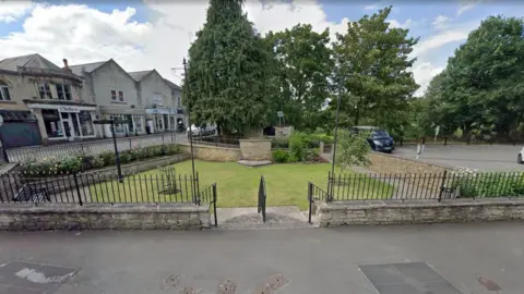 Google Queen Mary Garden. It is a triangular shaped grassy patch in the middle of a town square. There is a stone wall and iron railings around its circumference and several shops to its left and a carpark to its right. 