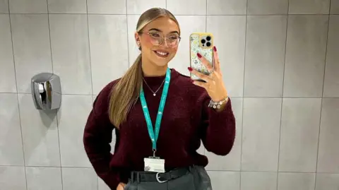 Lily-May Edwards She wears a burgandy jumper tucked into her grey trousers. Her hair is in a low ponytail. She has red nails and is taking a mirror selfie