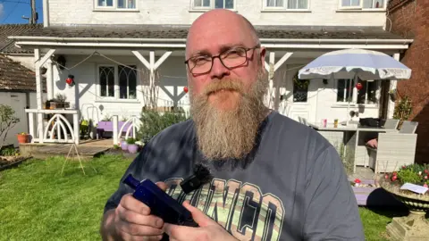 A man wearing a grey T-shirt, glasses and with a long beard, holding a blue victorian poison bottle 