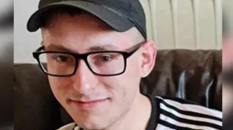 Essex Police Martyn Richardson, wearing glasses and a cap, looking at the camera. He is sitting on a sofa and is wearing a black and white top.