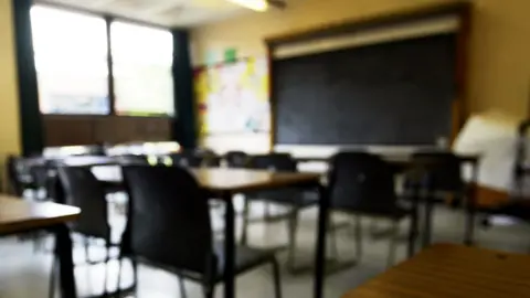 A blurred image of a generic classroom. A blackboard is situated at the far wall, in front of which sit rows of single-person desks with black chairs. 
