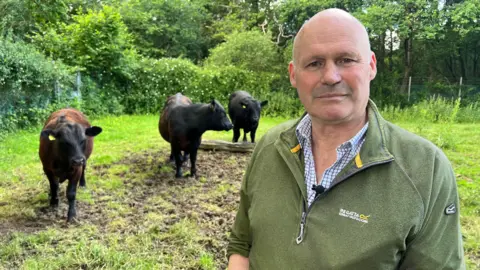 BBC/Ben Leete A bald man wearing a green jumper stands in a field. There are three black cows in the background. 