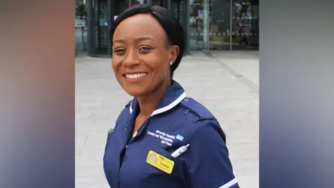 Leona is standing outside the main entrance of Coventry's University Hospital. She has her left shoulder pointing to the camera and is wearing her blue nurses uniform with white trimming.