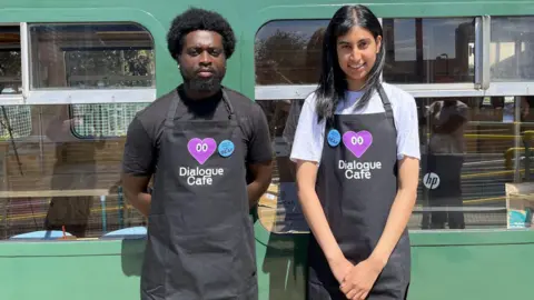 Two young baristas, wearing black aprons with the Dialogue cafe logo on the front - which includes text in white and a large purple heart - stand in front of the café's windows.