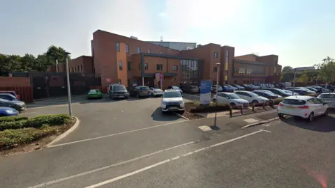 Google A Google Streetview image from the hospital, a modern brick and glass building,  with cars parked outside. 