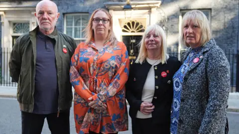 Steve Kelly, who has a bald head, white stubble and a green jacket, Charlotte Hennessy, with light ginger hair and wearing an orange floral dress, Margaret Aspinall, with shoulder length blonde hair and a black jacket, and Sue Roberts, who has blonde hair and a grey and black woollen coat, all stand looking into the camera with determined expressions with the black door of Number 10 Downing Street in the backdrop.