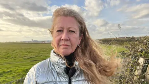 A woman with ginger hair and silver coat is standing next to Felton Common, a large green space