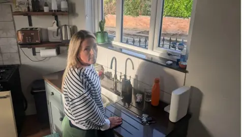 Alex Moss/BBC A woman standing near a kitchen window. She has blonde, shoulder-length hair and is wearing a black and white striped jumper and green leggings. She is stood in front of the sink area. Out of the window, a cobbled road can be seen with large slabs missing from the pavement, leaving bare patches of earth and uneven gaps. 