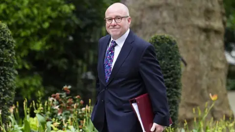 PA Media A man wearing a dark blue suit and tie with a geometric pattern in blues, pinks and purples, carries a dark-red ring-binder file with paper inside. He smiles at the camera as he passes. He is wearing glasses and has stubble.