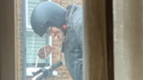 An older man in a black motorcycle helmet and leather jacket has a cigarette in his mouth as he stands beside a motorbike.