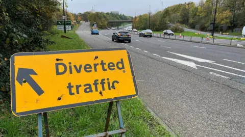 A yellow sign diverting traffic off the central motorway in Newcastle. Two cars are leaving the road and heading up a slip road. Rose Bridge can be seen in the distance across the motorway.