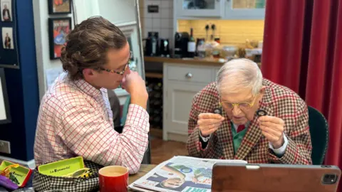 Jack Lupton A young man in a white shirt sits beside David Hockney, a man wearing a tweed jacket who is trying on a pair of new glasses in front of a newspaper.