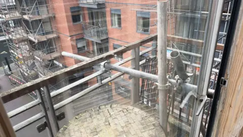 A view looking out of an apartment block window towards the ground. Outside you can see lots of metal scaffolding directly in front of the glass, as well as in front of other windows in the building opposite.
