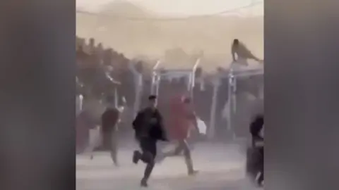 A crowd of people, one climbing over a fence, run to collect aid