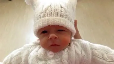 A baby in a knitted white cardigan and matching hat, being held