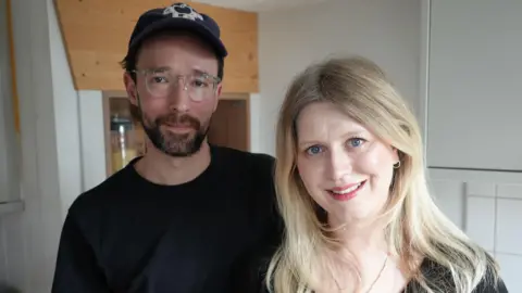 Qays Najm/BBC David and Hannah Weller, standing side by side, both looking down the camera. They are inside their kitchen, with a glimpse of tiles and a cupboard behind them. David is slightly taller than Hannah and is standing on the left of the image. He is wearing a black top and a dark blue cap, with a white logo on the front. He has a short, dark haired beard and wears glasses with clear plastic frames. Hannah is also in a black top. Her long blonde hair, falls just below her shoulders. She is smiling and showing her teeth.