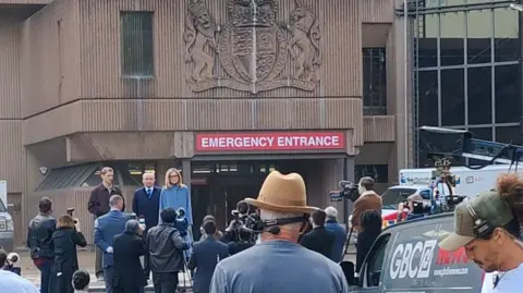 Out In Liverpool Film crew are stood outside Liverpool Crown Court which is being used as a film set. A red sign across the front of the building says Emergency Entrance in white capital letters. A large amount of film crew are gathered as filming takes place with actors stood on the steps.