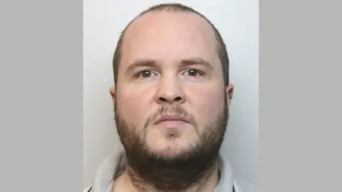 A custody picture of Matthew Ely. He has a dark scruffy beard and thinning dark brown hair on his head. He has small silver-coloured piercings in his left ear and just below his mouth