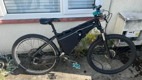 Wiltshire Police A black e-bike leant up against a windowsill outside a home, with blue handlebars and pedals.
