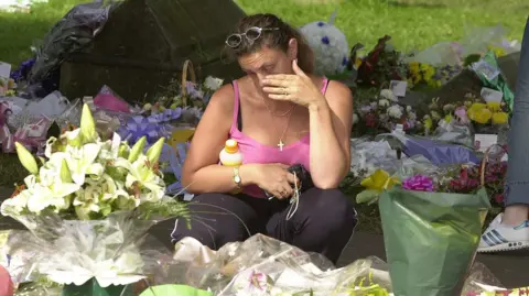PA Media A woman wearing a pink vest top crouches down among lots of flowers laid in memory of Holly and Jessica. She is wiping a tear from her eye.