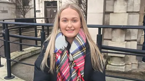 Leah McIlwrath has long blond hair parted in the middle and is wearing a dark coat and tartan scarf