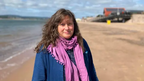 BBC Nicky Nicholls stood on Exmouth beach. She has long brown hair draped past her shoulders, is wearing a blue jacket and a pink scarf.