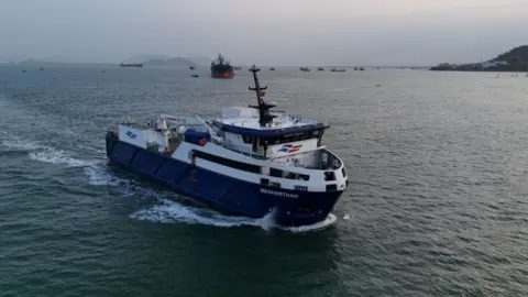 Isles of Scilly Steamship Group A blue and white freight vessel travels along  the surface of calm waters. The Menawethan logo is printed on the side of the boat.