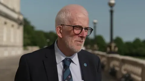 Stewart Dickson standing outside Stormont. He is wearing black-framed glasses, a black suit jacket, a striped tie and white shirt.