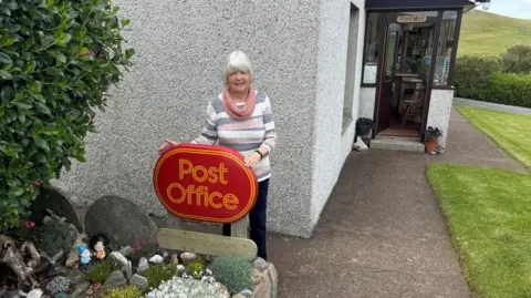Woman with white hair and stripy jumper stands next to post office sign. Behind her is a house and a porch. 