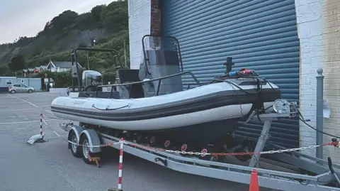 Round Britain eRIB A grey electric powered inflatable boat moored up in front of metal shutters in a car park and fenced off with red and white chain