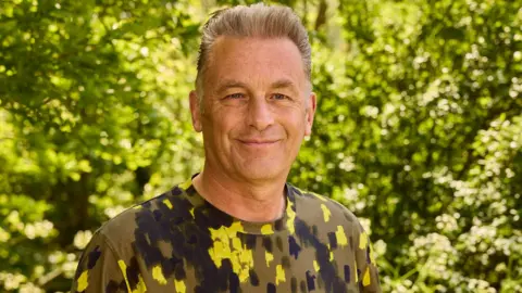 A man with spiky grey hair wearing a green camouflage t-shirt stands outside surrounded by green bushes in the sunshine