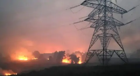 A wildfire with a smoky sky and an electricity pylon in the foreground