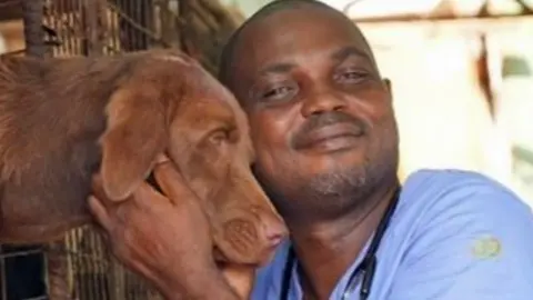 Dr Mark Afua, a vet in Lagos wearing blue scrubs and a stethoscope, hugs a brown dog whose head is poking out of a metal cage.