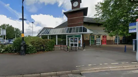 Exterior view of a Morrisons supermarket building with a clock tower above the main entrance. The image shows the front of the store, a covered walkway, railings, hedges and part of the car park in the foreground. Trees stand to the right of the building under a partly cloudy sky. 