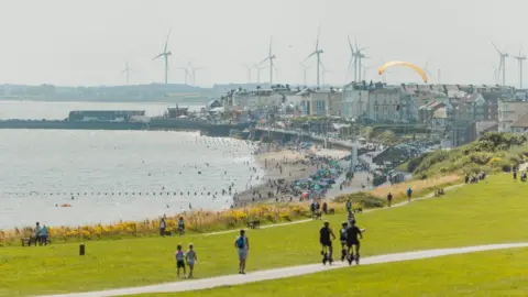 VHEY A view of the seafront at Bridlington. There is large stretch of beach leading to the sea, with rows of buildings in the background. There are several people making their way along a path, some on bicycles.