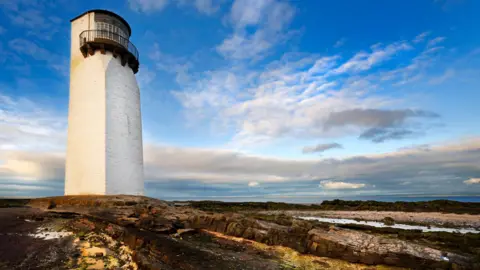 Getty Images An old white lighthouse stands on a rocky shoreline with water in the distance under a cloud-flecked blue sky