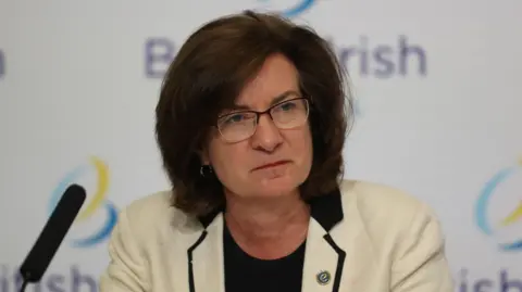 PA Media Eluned Morgan sat in front of a banner that says British Irish council, wearing a cream jacket and black top. She is wearing glasses.
