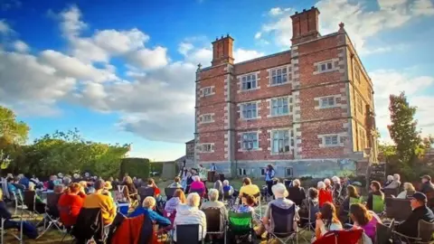 A large block-shaped brick building with four floors and large brick chimneys with a crowd of people sitting outside on camp chairs looking toward the building