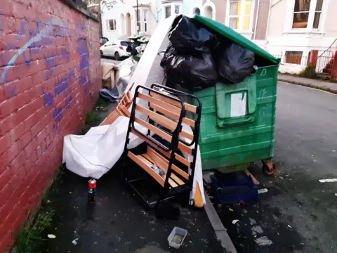 A large green bin with a bed frame and mattress propped against it. 